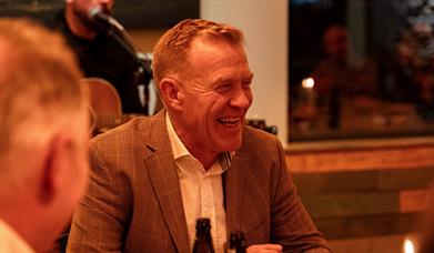 Adam Henson sits at a table in a dinner jacket smiling off to one side. A musician sits behind him with a microphone.