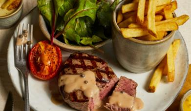A seared steak with peppercorn sauce accompanied with grilled tomato, chips and green leaves